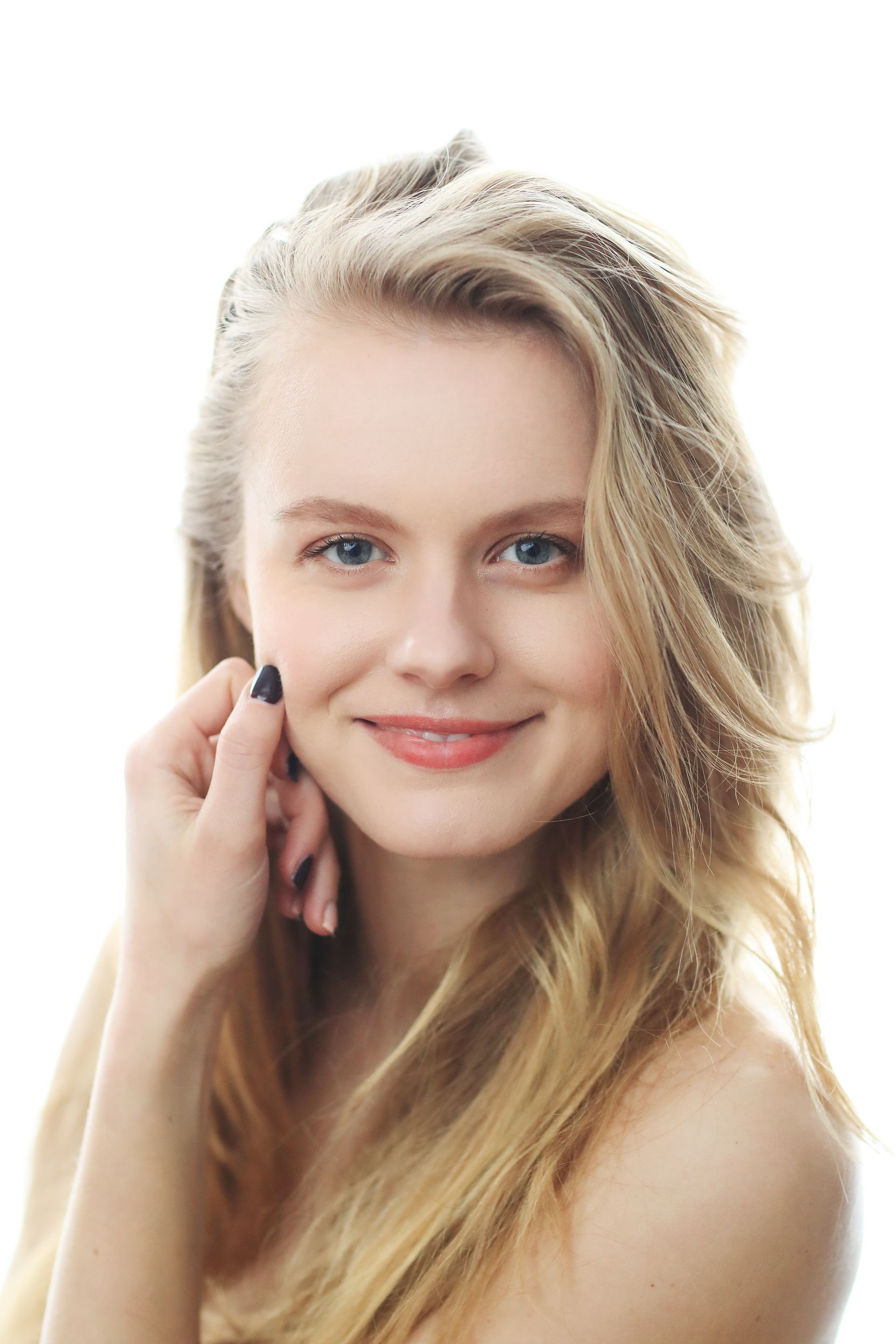 A woman with long blonde hair is smiling and touching her face.