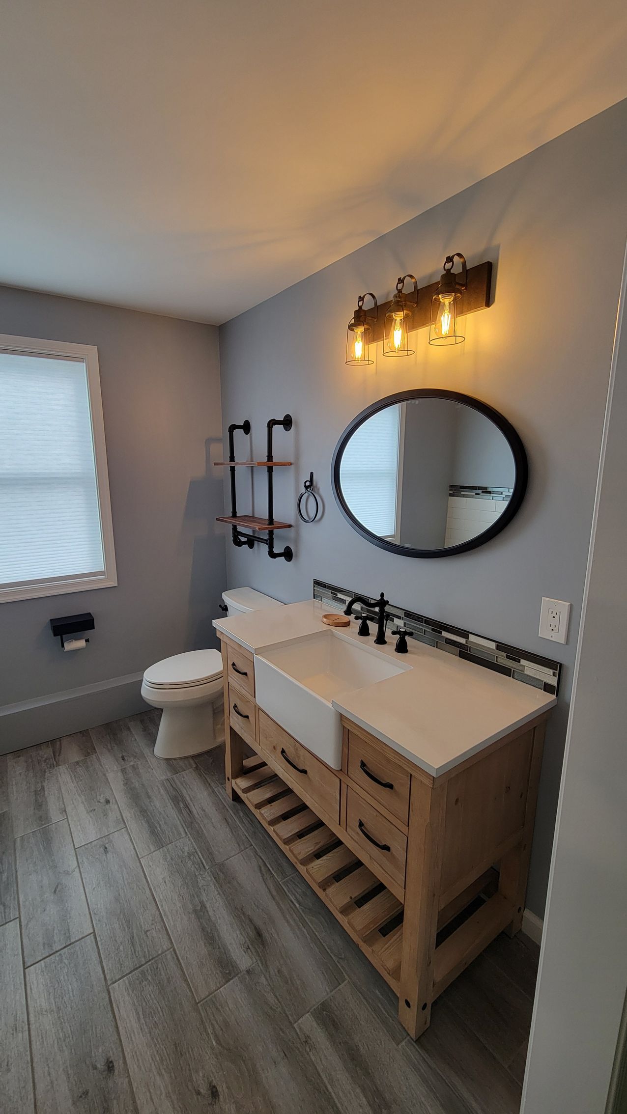 Bathroom with gray walls, wood vanity, and a black-framed mirror. Features industrial-style lighting and shelving.