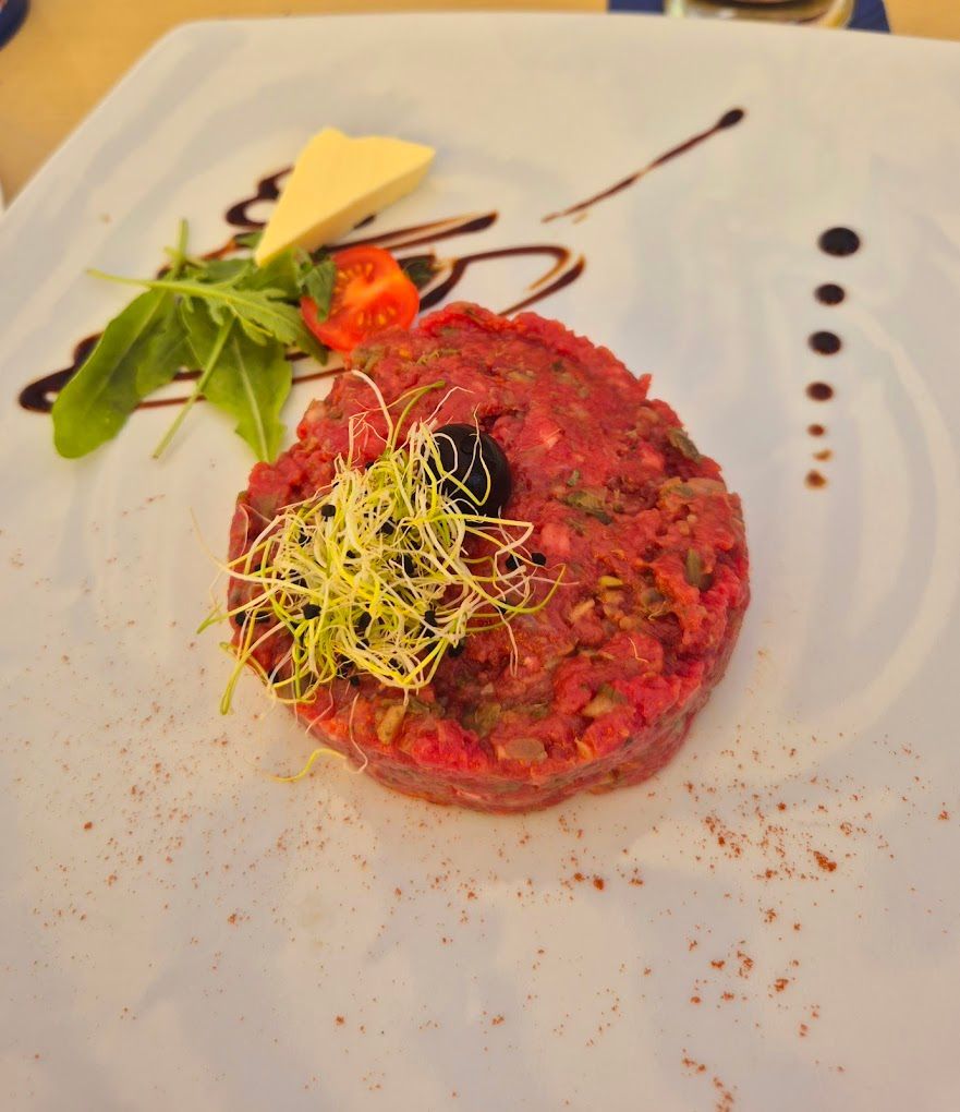 Steak tartare avec garnitures sur une assiette blanche; comprend du fromage, de la roquette, des tomates cerises et un filet de balsamique.