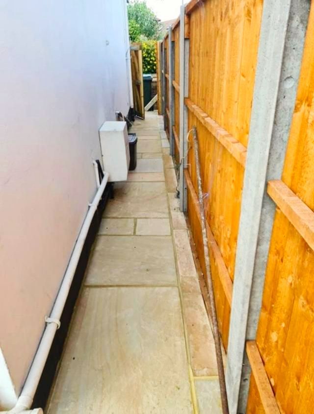 Narrow paved walkway between a pale pink wall and a wooden fence.
