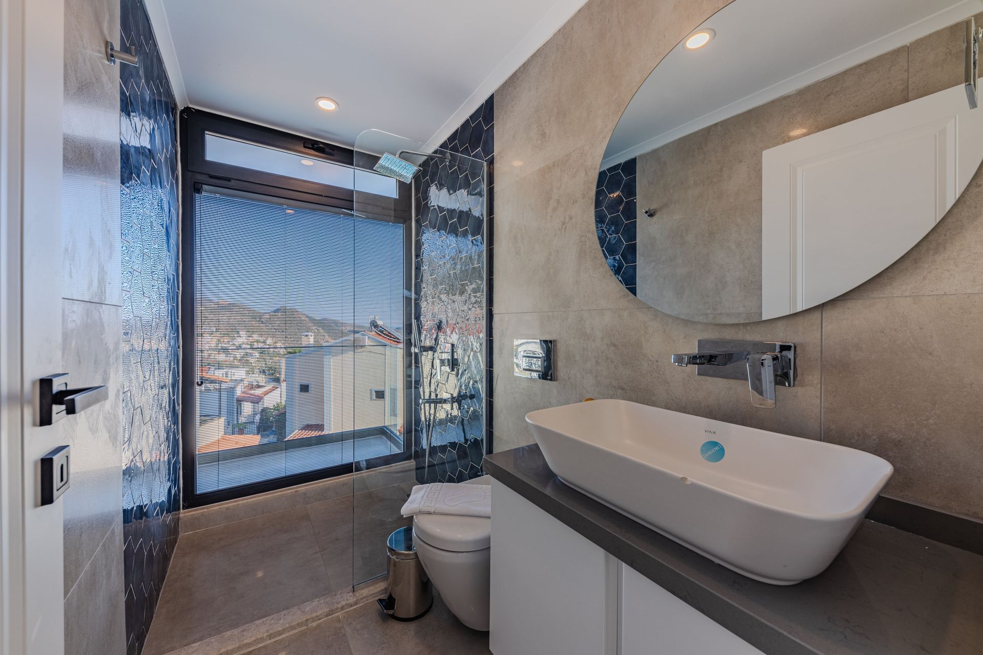 Modern bathroom with floating sink, round mirror, toilet, and large window overlooking a city view