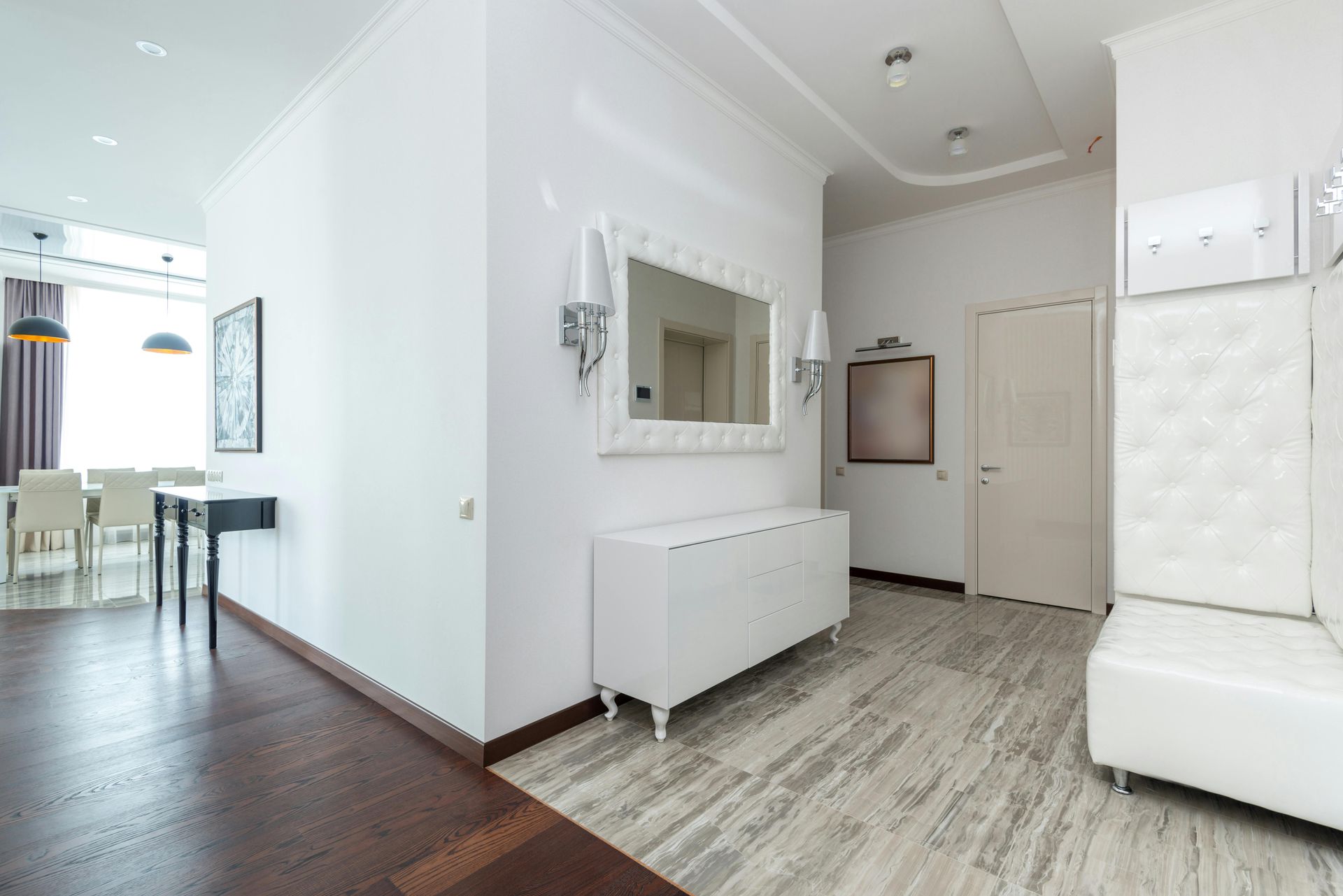 Bright modern entryway with white furniture, mirror, and light gray wood flooring adjoining a dining area