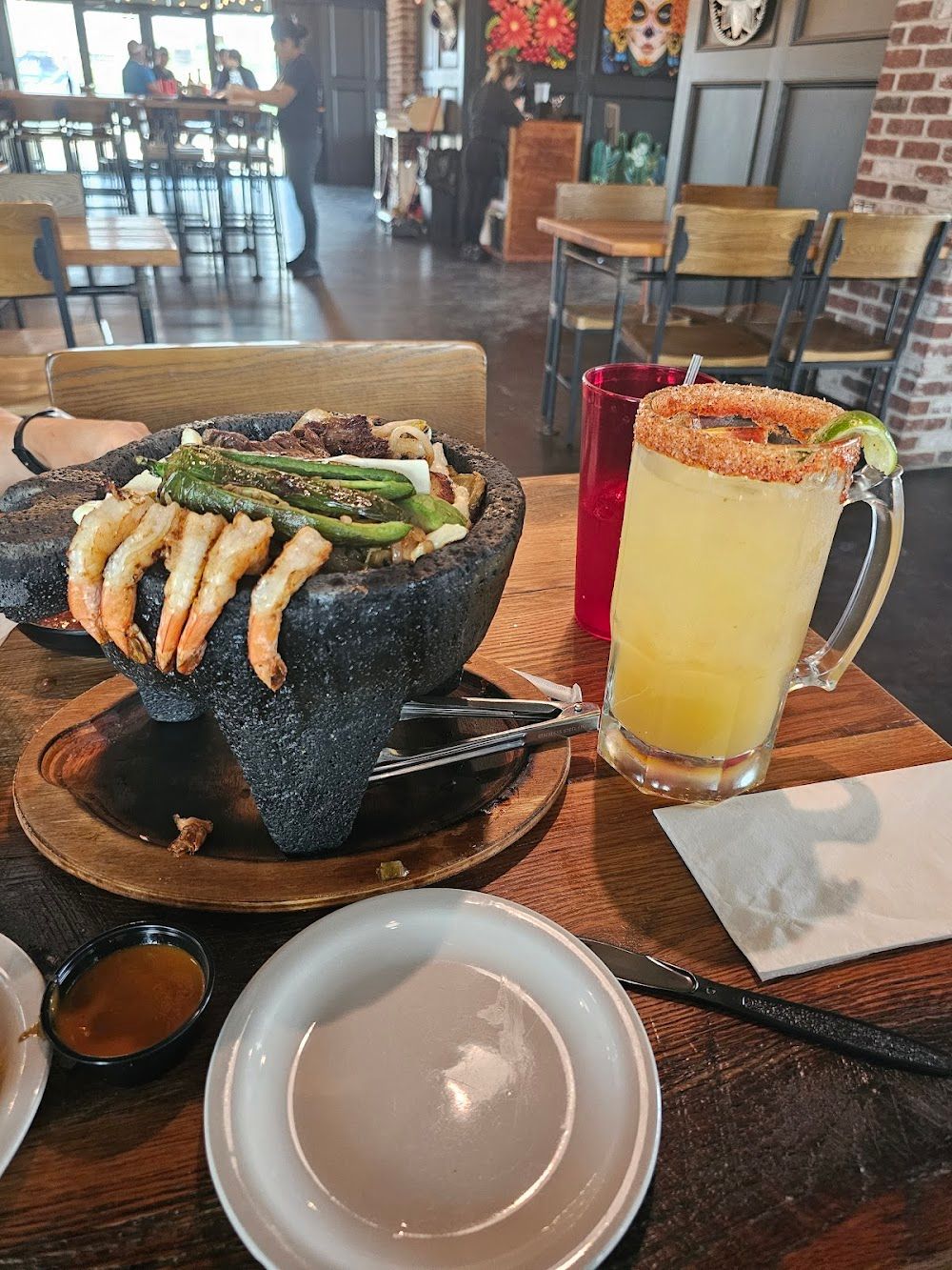 Black stone seafood dish with shrimp and green garnish beside a yellow cocktail on a restaurant table