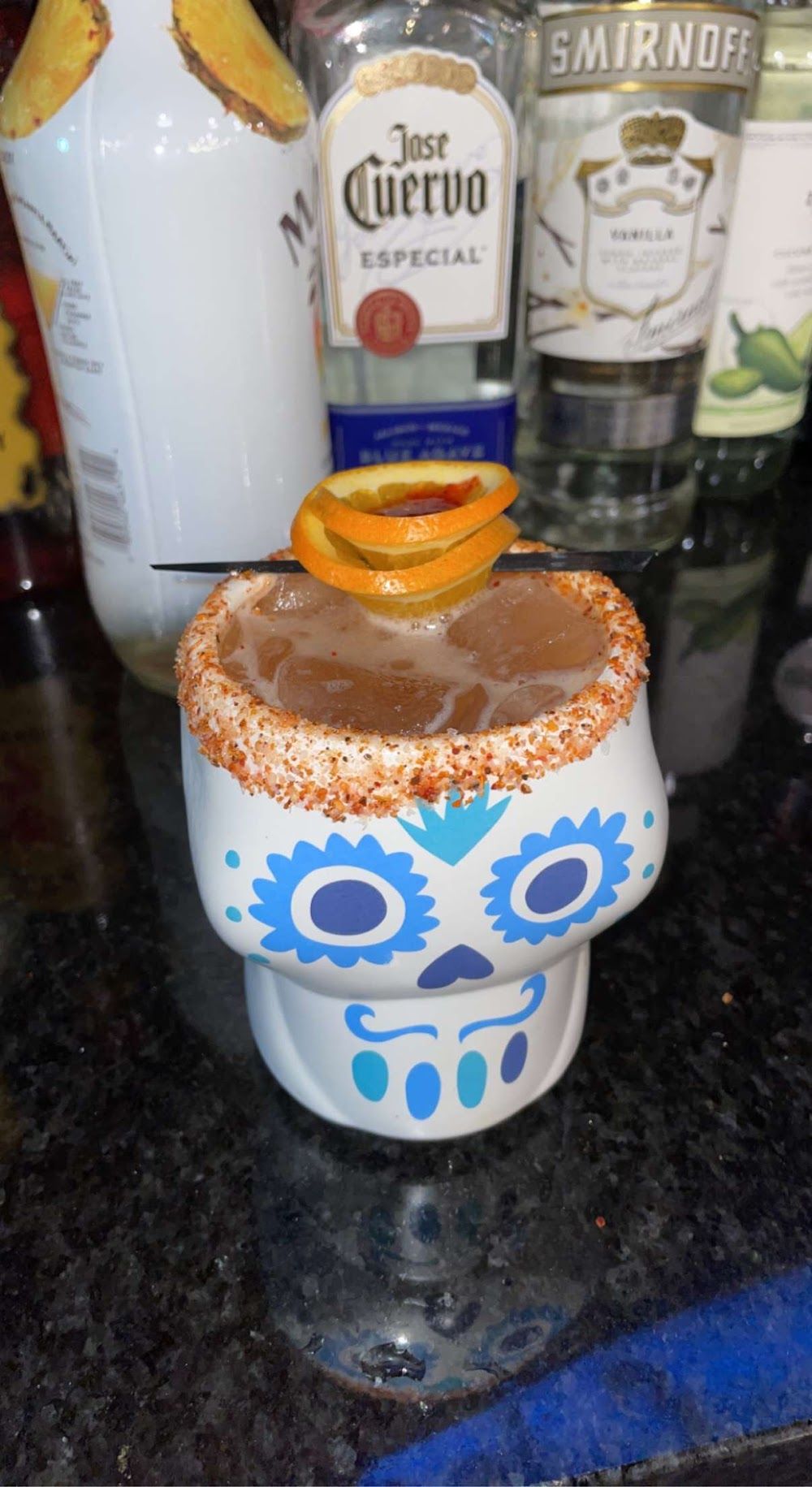 Decorative skull-shaped cup with orange drink and orange garnish on a countertop, in front of liquor bottles