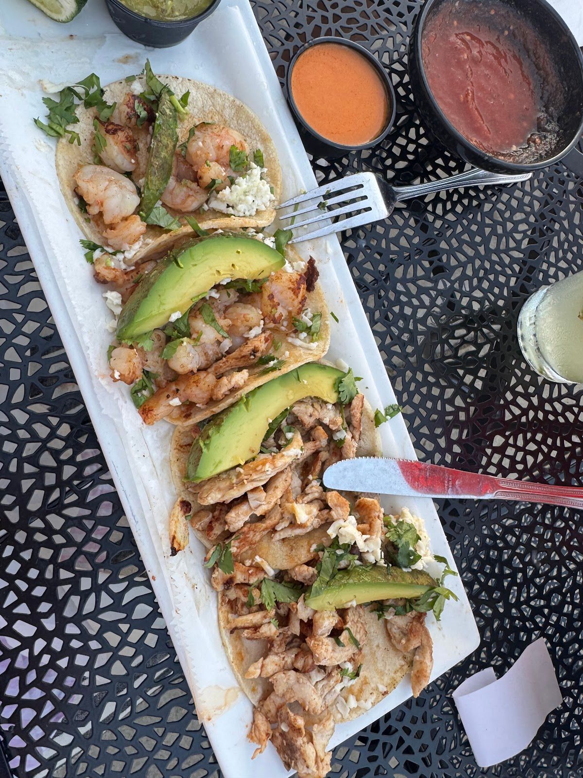 Tacos on a white tray with avocado slices and orange and red dipping sauces on a black table
