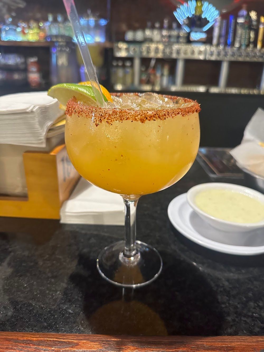 Orange cocktail with salted rim and lime wedge on a bar counter, with a bowl of dip in the background