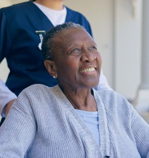 A smiling person in a grey cardigan looks forward, with a healthcare worker in blue scrubs standing behind them.
