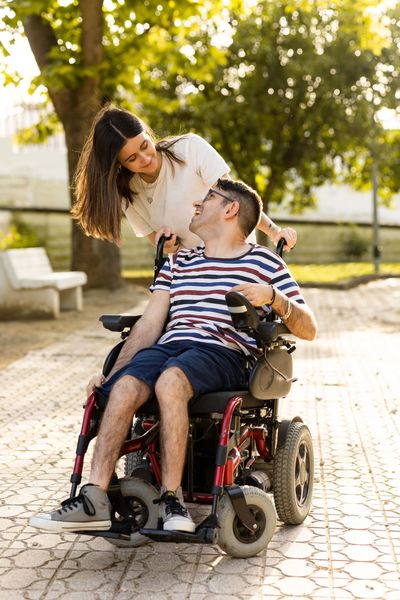 A woman leaning over and smiling at a man sitting in a motorized wheelchair in a park.