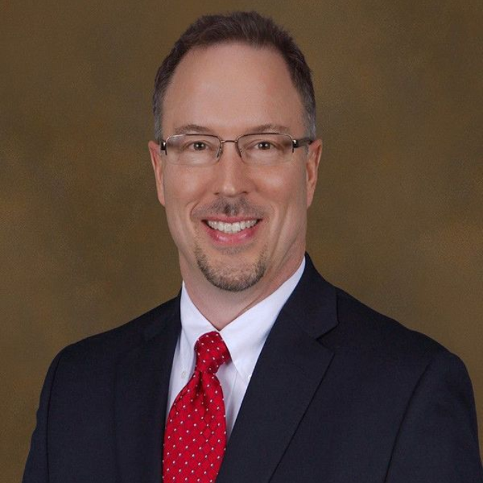 Man in a suit smiles, wearing glasses and a red polka-dot tie, against a brown background.