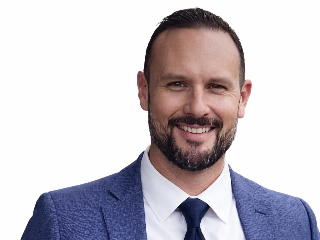 Man in blue blazer smiles, dark tie, short brown hair and beard, white background.