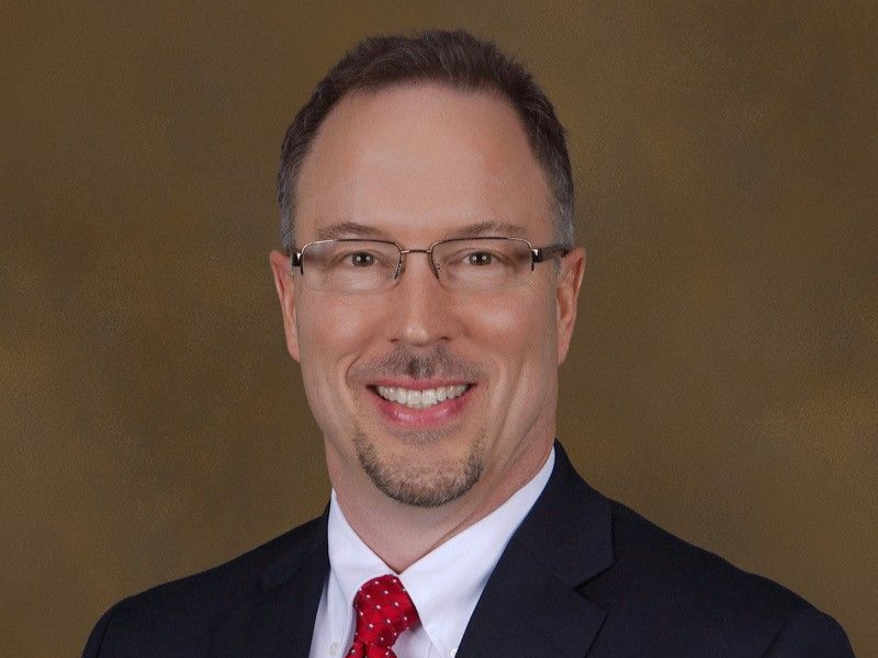 Man with glasses smiling, wearing a suit and red tie, in front of a neutral background.