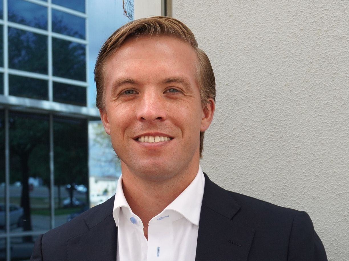 Man in a suit smiles outdoors near a building with blue windows.