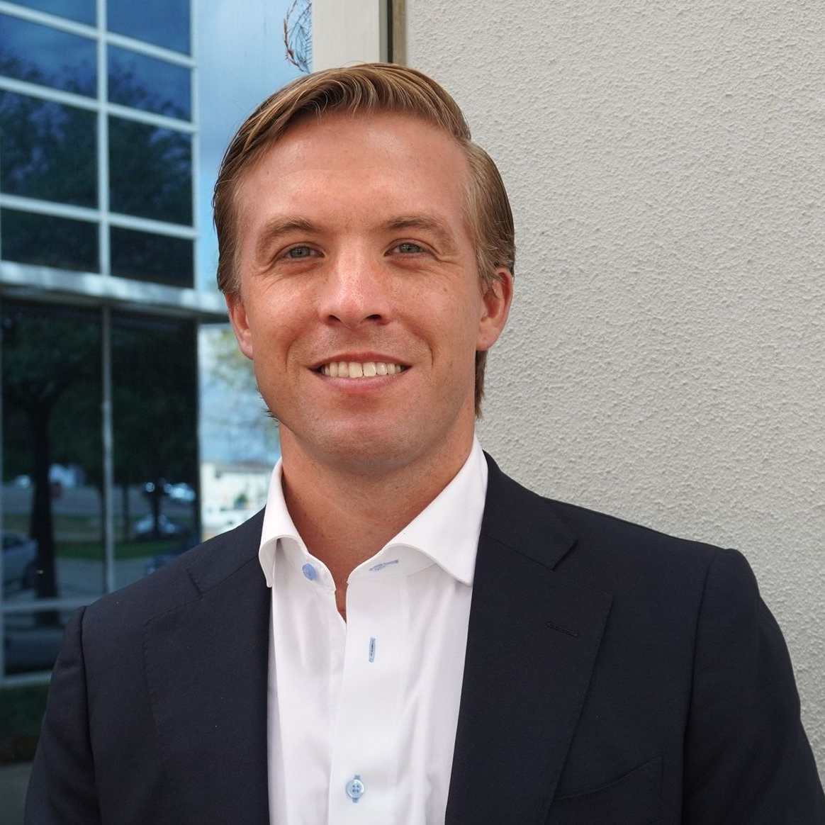 Man in a suit smiles outdoors, a building and windows in the background.