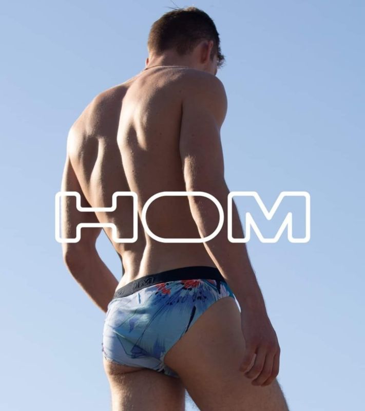 Uomo in slip da bagno fantasia, di spalle, contro un cielo azzurro. Logo HOM sovrapposto.