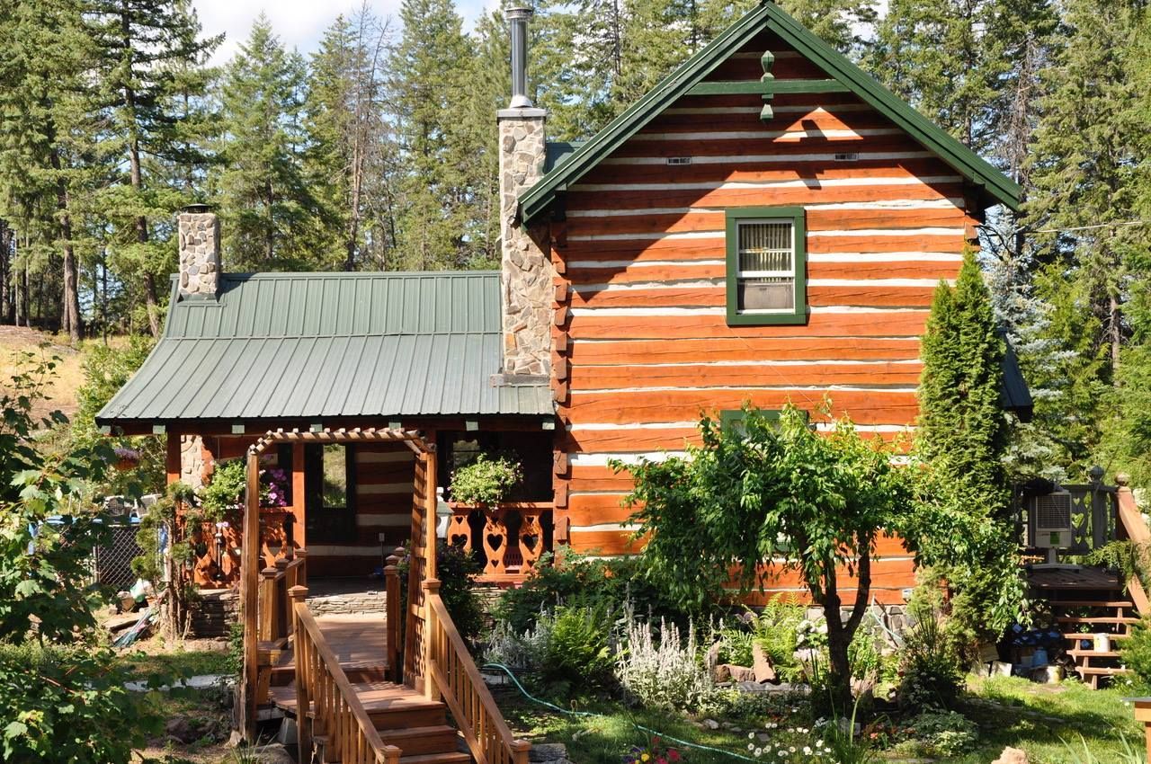 A two-story log cabin with a green metal roof, stone chimney, and wooden walkway surrounded by a forest.