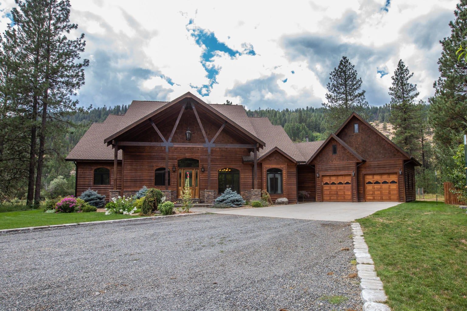 A brown wood-sided house with a steep roof and double garage, situated in a wooded area with a gravel driveway.