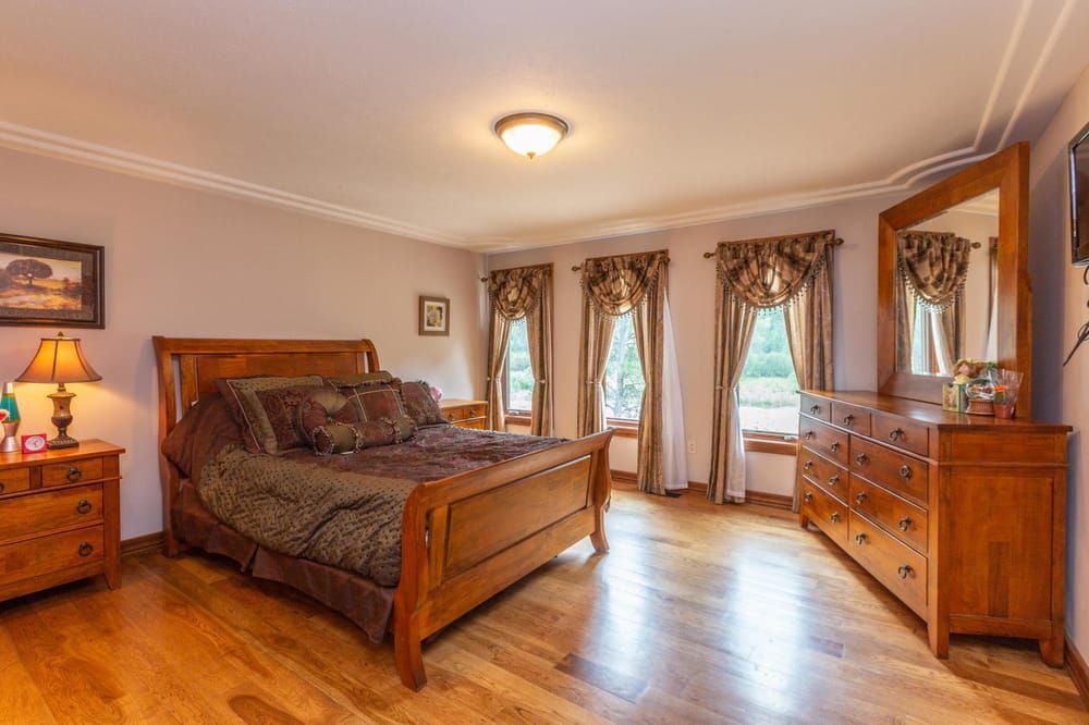 A bedroom with light walls, hardwood floors, a wooden bed set, a dresser with a mirror, and windows with ruffled curtains.