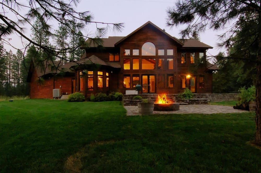 A large, two-story wooden house at twilight with a glowing fire pit on a stone patio in the backyard.