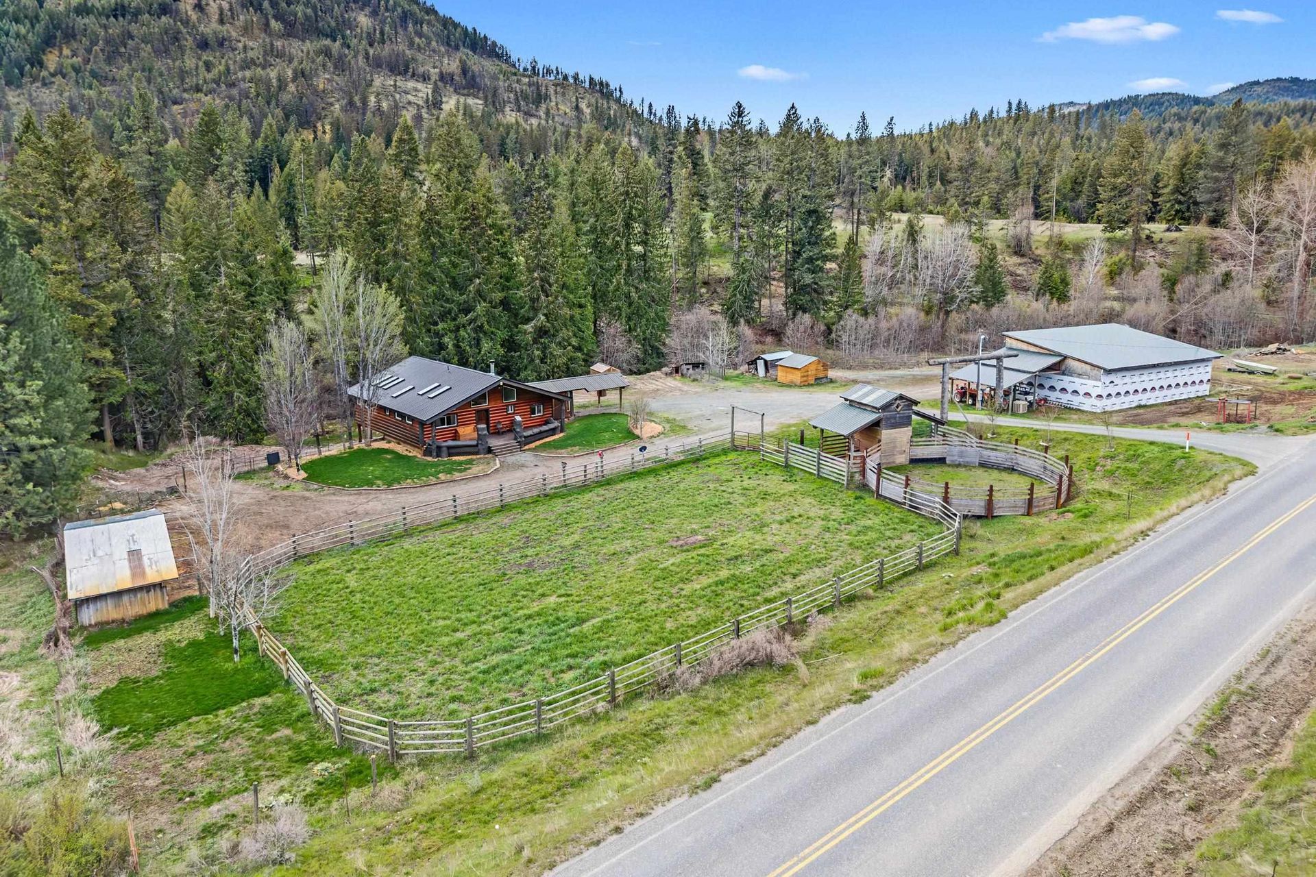 Rural homestead with house, outbuildings, fenced green field, and road beside forested hills