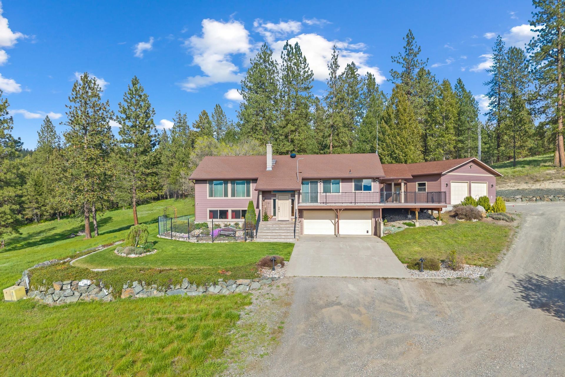 Suburban house with brown roof, garage, and gravel driveway on a grassy hillside under a blue sky.