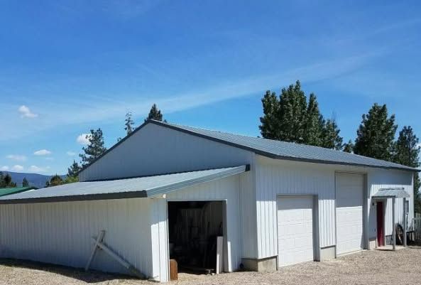 A white metal-sided workshop with a gray roof and two closed garage doors set against a blue sky with pine trees.