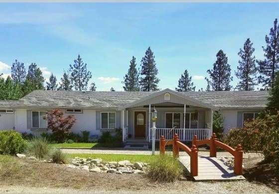 A white, single-story house featuring a front porch with a red decorative bridge leading to the entrance under tall trees.