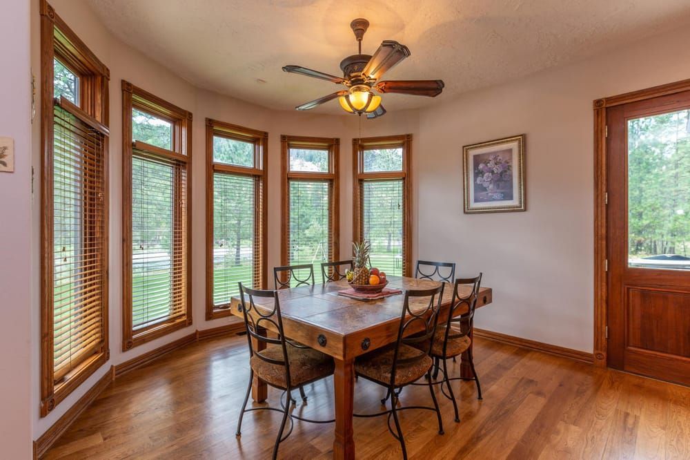 A sunlit dining room with a wooden table, chairs, large windows, and a ceiling fan on hardwood floors.