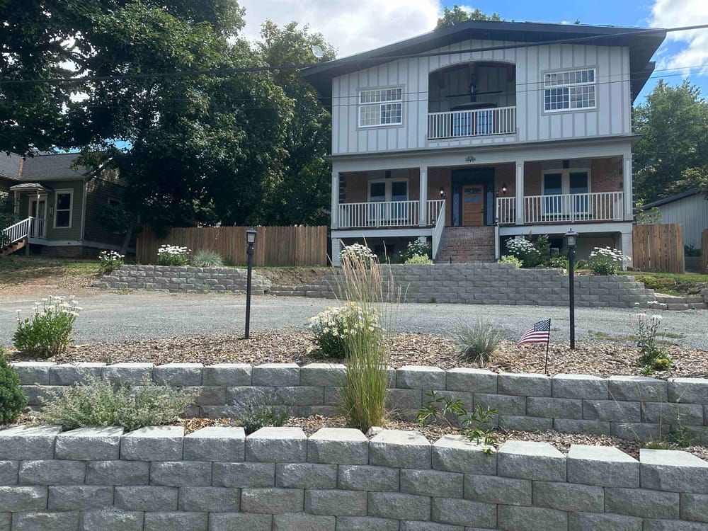 A two-story white house with a porch sits behind multiple grey stone retaining walls and a gravel yard.