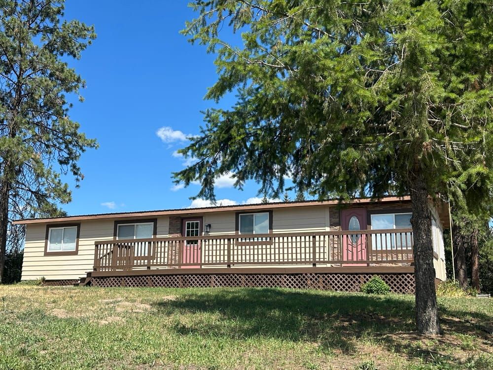 A tan, single-story home with a long wooden deck, set against a bright blue sky and surrounded by evergreen trees.