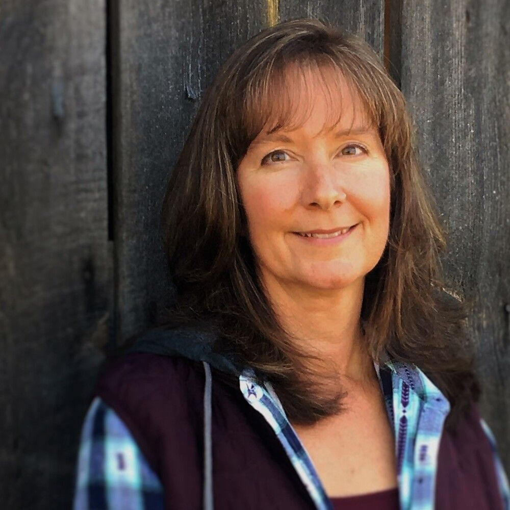 A person with long brown hair, wearing a blue plaid shirt and dark vest, smiling against a dark wooden wall.