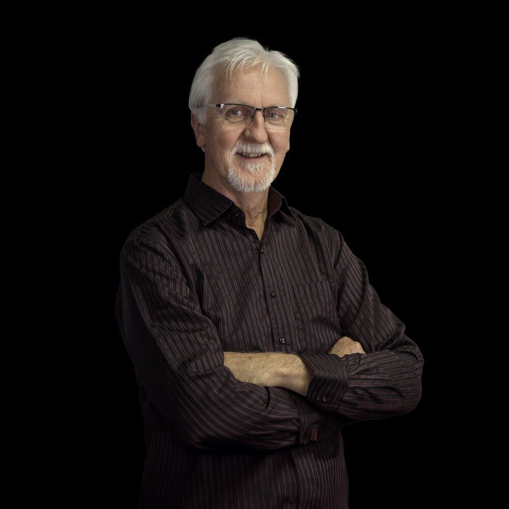 A man with white hair, glasses, and a goatee, wearing a patterned button-down shirt, stands with arms crossed on black.