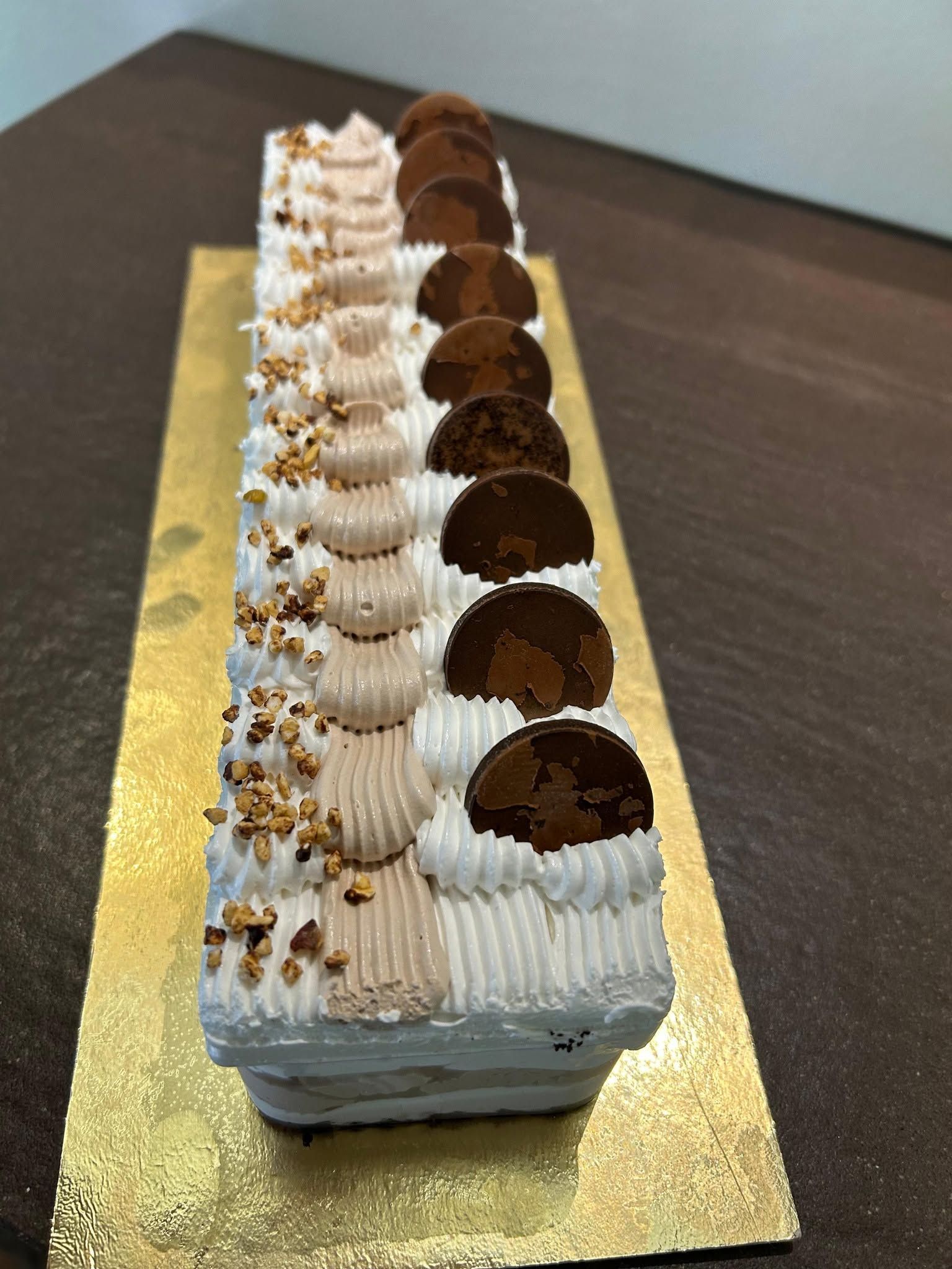 Torta rettangolare a strati, ricoperta di glassa bianca e decorazioni al cioccolato, su una base dorata.