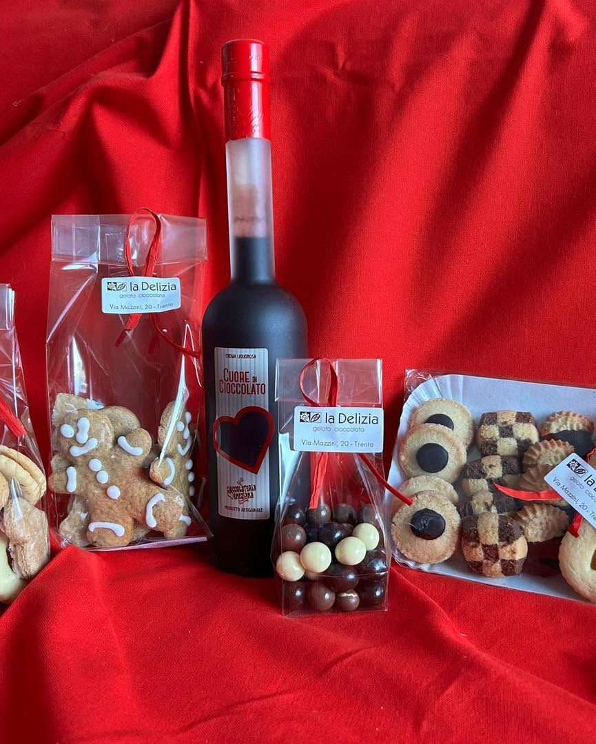 Assortimento di biscotti e dolciumi confezionati, disposti intorno a una bottiglia di vino rosso su uno sfondo di stoffa rossa.