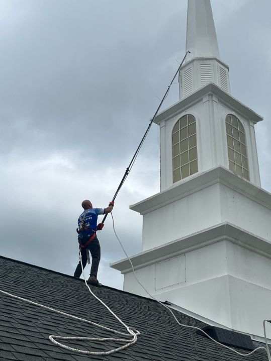 Man on a roof cleaning a church steeple with an extended cleaning pole. Cloudy sky.