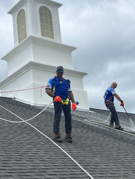 Two workers on a roof, attached to safety ropes, cleaning the surface with sprayers, with a white steeple in the background.
