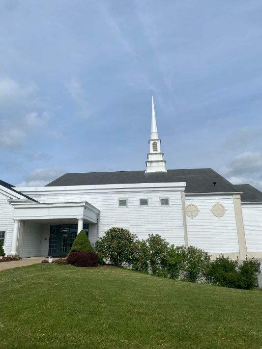 White church building with steeple, on a green hill, under a cloudy sky.