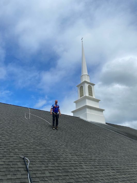 Person on a shingled roof, church steeple in the background under a cloudy sky.