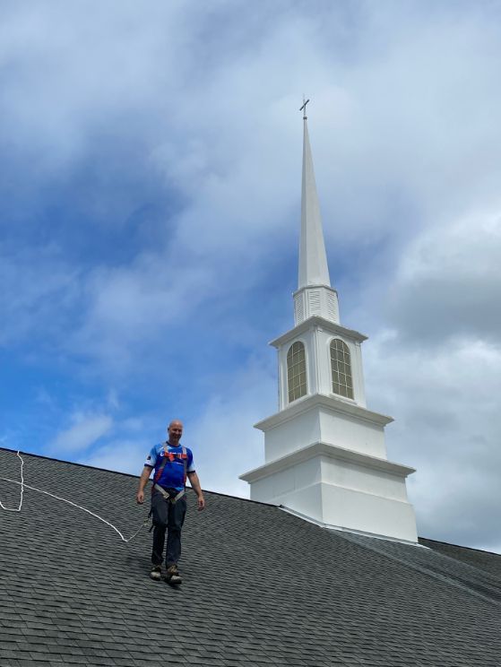 Person in safety gear on a dark shingle roof, near a white church steeple against a cloudy sky.