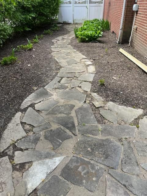 Stone path winds through a garden, past a fence and a brick building.