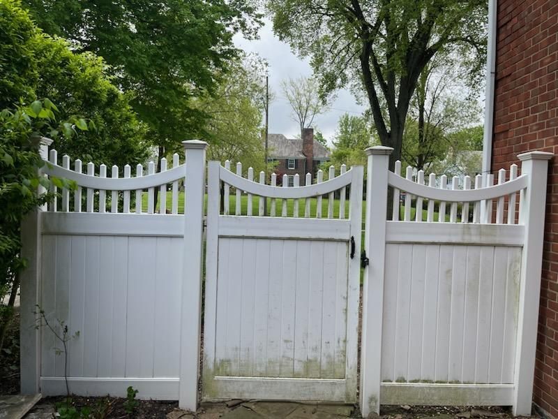 White picket fence with gate, set against trees and a brick building, shows slight discoloration.