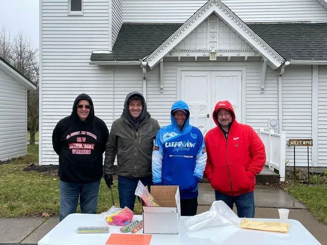 Four people stand behind a table outside a white church. They wear hooded jackets. A box and other items are on the table.