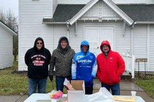 Four people stand behind a table outside a white church. They wear winter jackets, and some have hoods up.