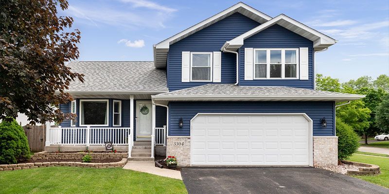 Blue two-story house with white trim, a two-car garage, and a porch. Green lawn with trees and a blue sky.