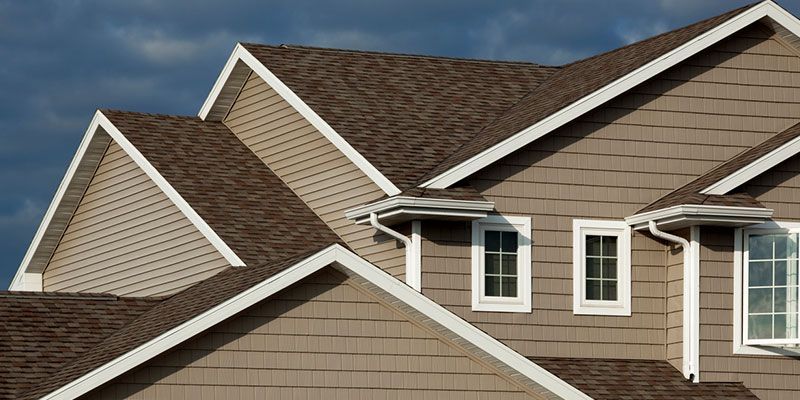 Close-up of a house with a brown roof and tan siding, white trim around the roof and windows.