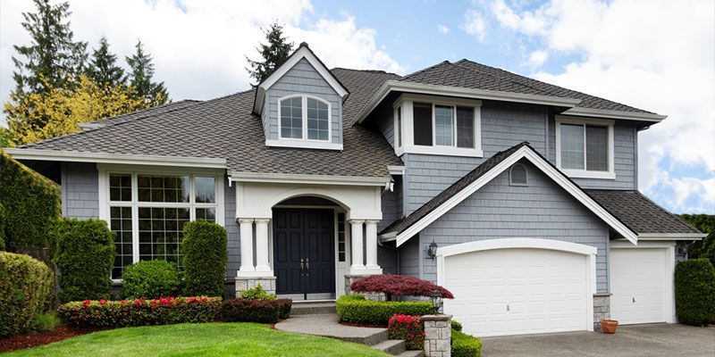 Gray house with white trim, dark blue door, two-car garage, and well-kept landscaping under a cloudy sky.