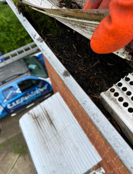 A person wearing an orange glove cleans a gutter filled with debris. A