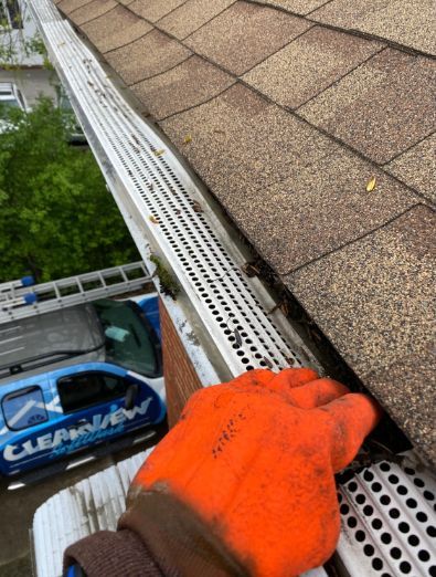 Person in orange glove cleaning a gutter covered with a screen on a roof near a parked van.