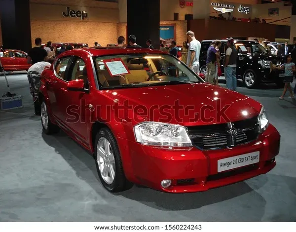 Dark blue sedan parked on a concrete surface, with a chain link fence and buildings in the background.