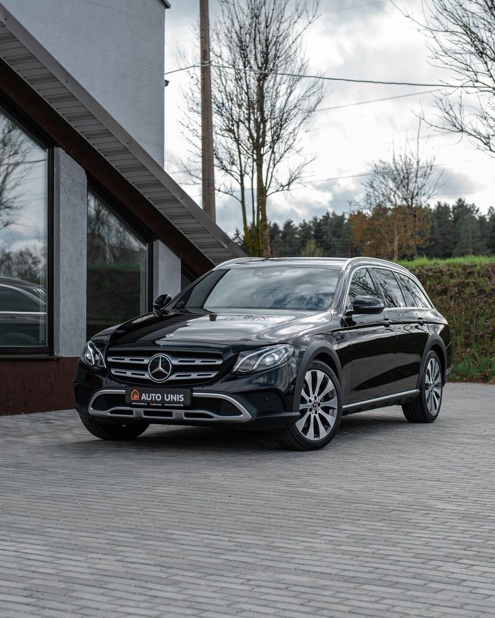 Black Mercedes-Benz E-Class All-Terrain parked on a cobblestone driveway in front of a modern building.