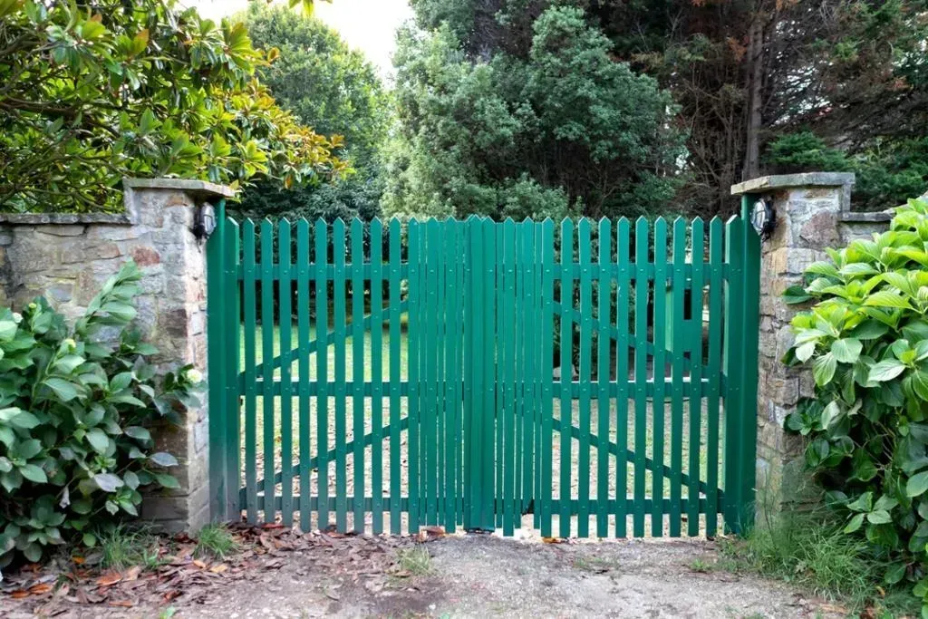 Green wooden gate in a stone wall, leading to a yard with green foliage.